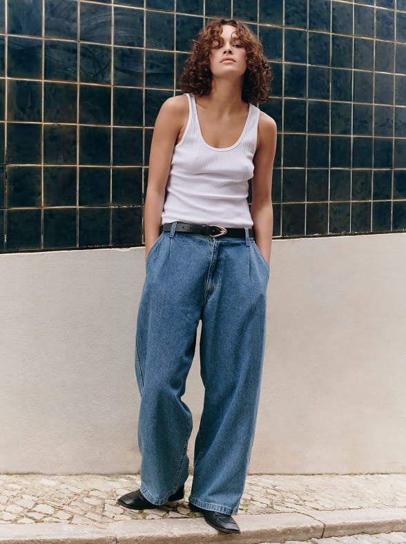 Woman in a white sleeveless top and beige pants standing on a sunlit stone pavement with an arched window in the background
