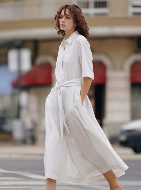 Woman in a white patterned dress standing outdoors in a sunlit street with a pink building in the background.