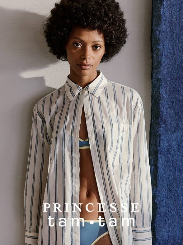 Woman in a white sleeveless top and beige pants standing on a sunlit stone pavement with an arched window in the background