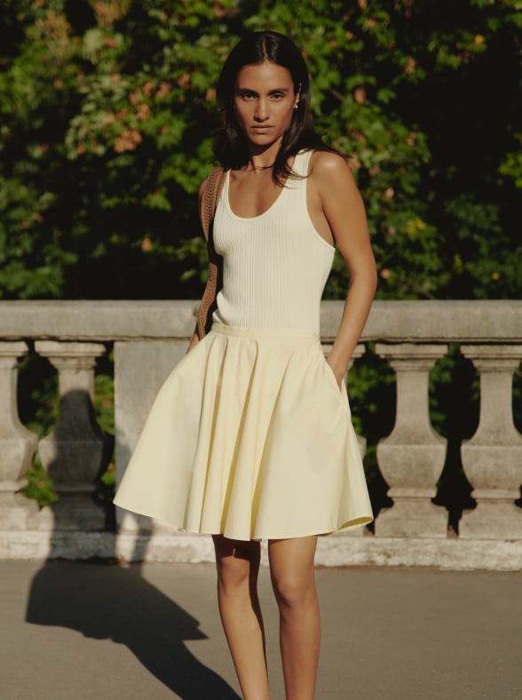 Woman in a white sleeveless top and beige pants standing on a sunlit stone pavement with an arched window in the background