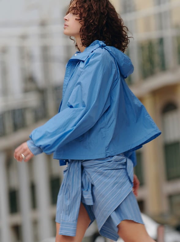 Woman in a white sleeveless top and beige pants standing on a sunlit stone pavement with an arched window in the background