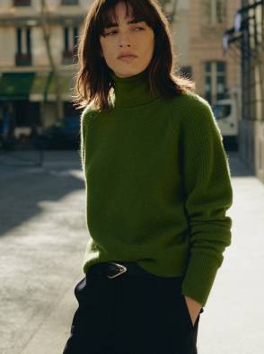 Woman in a white patterned dress standing outdoors in a sunlit street with a pink building in the background.