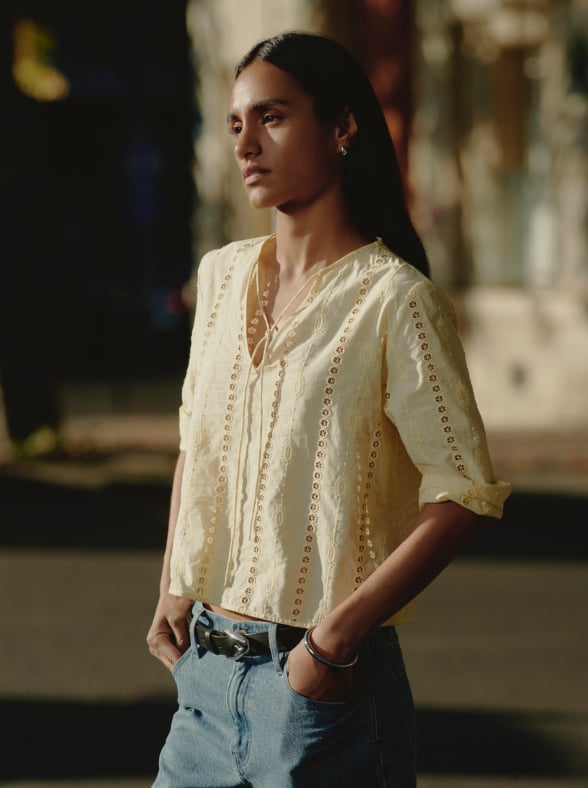 Woman in a white sleeveless top and beige pants standing on a sunlit stone pavement with an arched window in the background