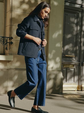Woman in a white patterned dress standing outdoors in a sunlit street with a pink building in the background.