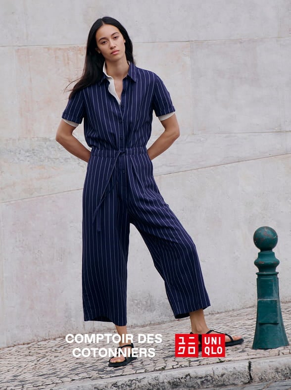 Woman in a white patterned dress standing outdoors in a sunlit street with a pink building in the background.